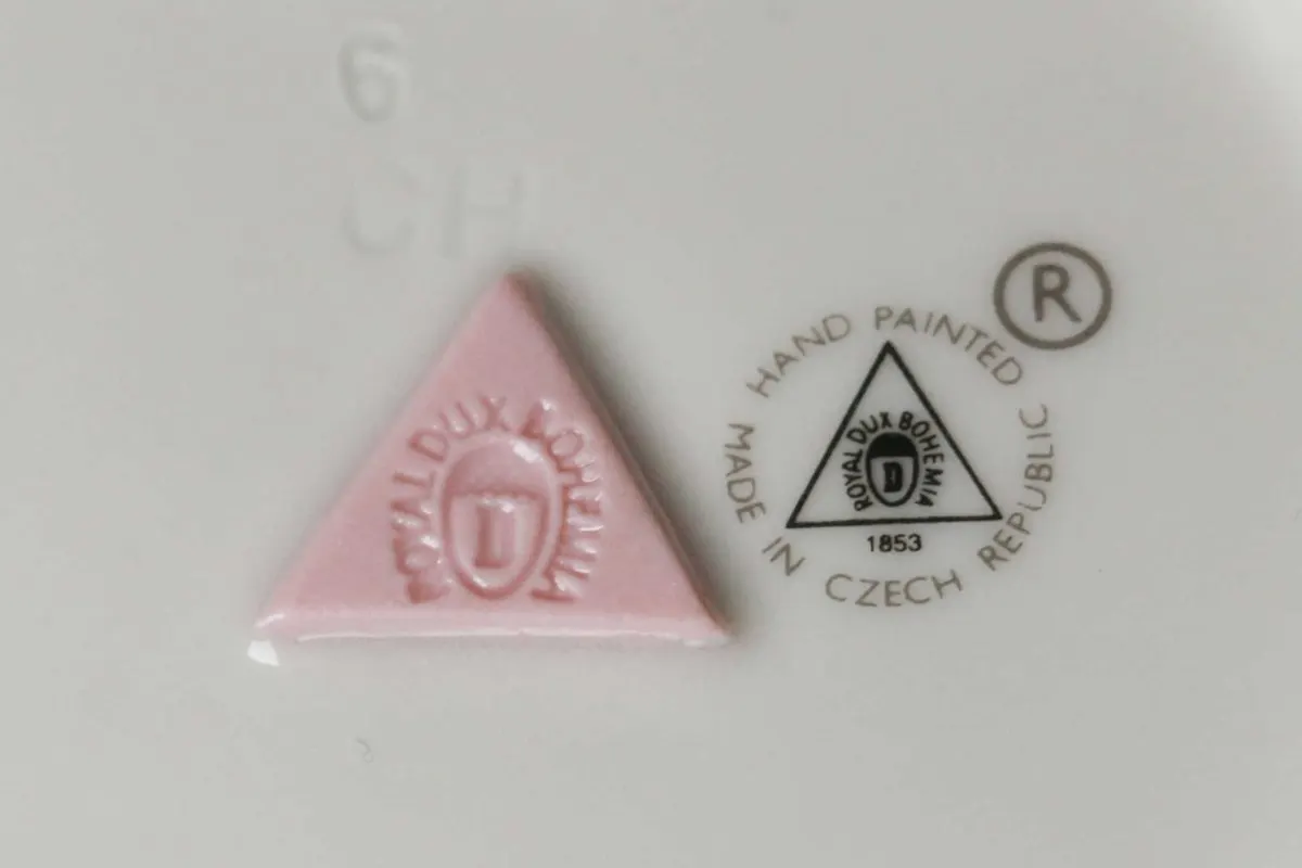 Plastisch gestaltete Marken sind in der Regel aufgeklebte Stücke der Porzellanmasse mit eingepresster Markierung. Bis heute werden sie z.B. von der Porzellanfabrik Royal Dux (Duchcov) verwendet – hier ein aufgeklebtes rot-gefärbtes Stück Porzellanmasse mit dem eingeprägten Namen der Manufaktur.
